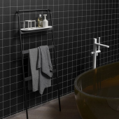 Mobile da bagno MrWOLF di Antonio Lupi in un ambiente contemporaneo con pareti di piastrelle nere, accanto a un lavabo in vetro marrone trasparente e rubinetteria moderna, decorato con una mensola metallica nera con prodotti da bagno e asciugamani grigi