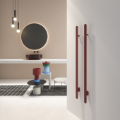 Scaldasalviette Rigo in ottone di Antonio Lupi posizionato verticalmente su una parete bianca con design minimale, accanto a un tavolino con oggetti di design e una lampada pendente in una stanza moderna e ben illuminata.