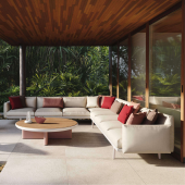 Elegante composizione del Divano Componibile Boma di Kettal in un lussuoso patio coperto, con cuscini rossi e beige, accanto a un tavolino rotondo in legno e metallo, immerso in un ambiente tropicale