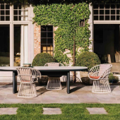 Spazio esterno elegantemente arredato con un set da pranzo KETTAL 'sedia cala' che presenta sedie intrecciate uniche e un tavolo nero elegante, ambientato su uno sfondo di giardino lussureggiante e pareti ricoperte di edera