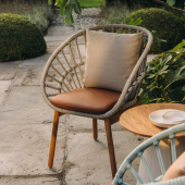 Vista ravvicinata di una sedia KETTAL 'sedia cala' con schienale intrecciato, seduta in pelle color cuoio e cuscino neutro, posizionata su un patio in pietra con vegetazione sullo sfondo."