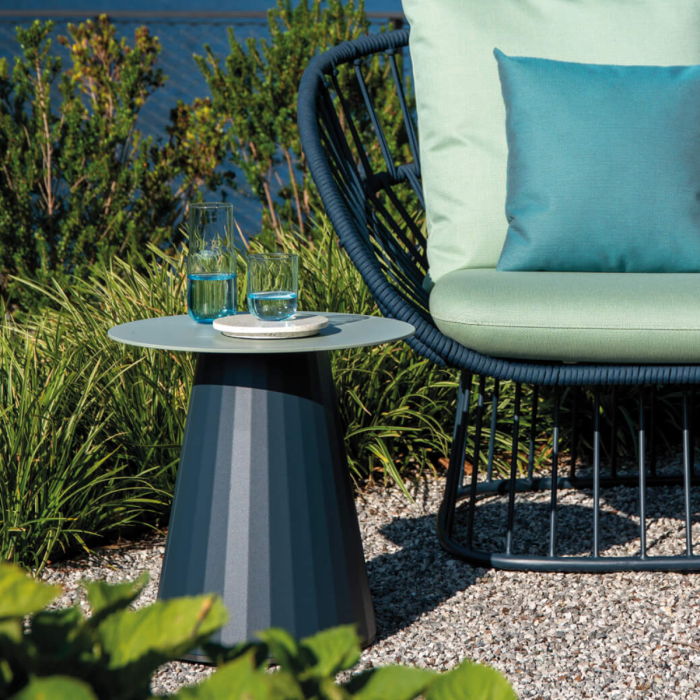 Tavolino Cala di KETTAL in alluminio di alta qualità, design moderno di Doshi Levien, presentato in un giardino estivo accanto a una poltrona di lusso con cuscini verdi
