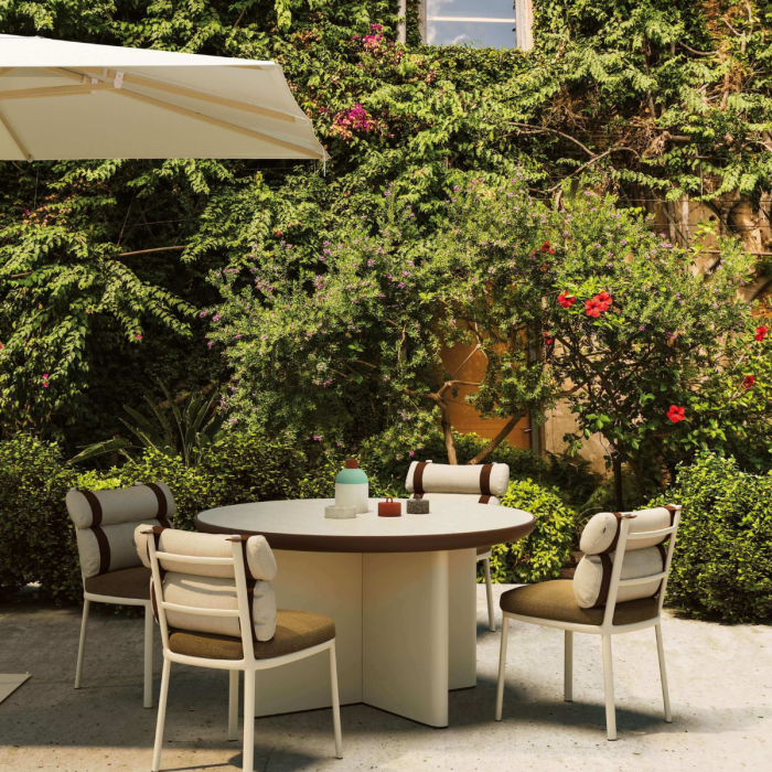 Tavolo Cala di KETTAL in un giardino ombreggiato, con sedie abbinate sotto un ombrellone bianco, immerso in un'ambientazione esterna fiorita."